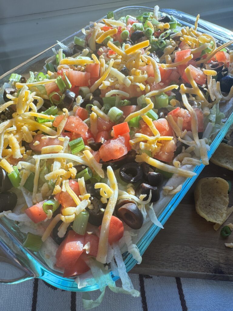 Taco dip with tomatoes, black olives, green onions and shredded cheddar cheese in a pyrex tray